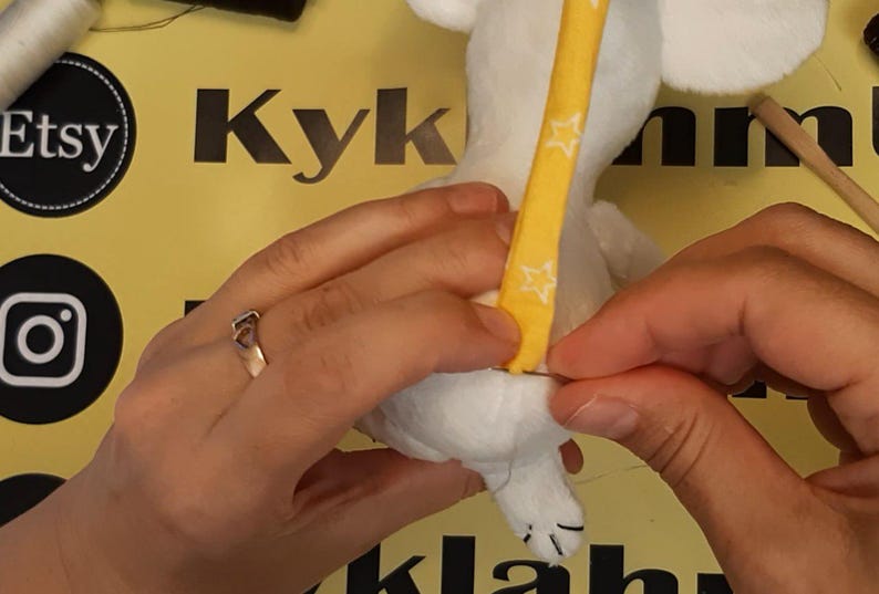 May include: A white plush toy being hand-stitched with a needle and thread. A yellow fabric strip featuring a white star pattern is being attached to the back of the plush toy. The toy has visible white ears and a small white tail with black stitching.