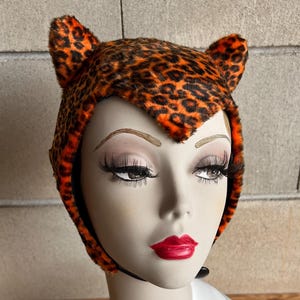 Könnte beinhalten: Ein Hut mit Leopardenmuster und ohrenförmigen Akzenten wird auf einem Schaufensterkopf präsentiert. Der Hut hat ein braunes und orangefarbenes Fleckenmuster. Die Schaufensterpuppe hat roten Lippenstift und lange Wimpern. Der Hintergrund ist eine hellgraue Wand und eine Holzoberfläche.