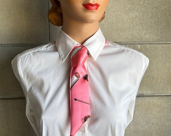 May include: A pink tie with a pattern of makeup items, including lipsticks and brushes, is displayed on a mannequin wearing a white collared shirt. The tie's design is set against a pink background. The mannequin has a vintage hairstyle and red lipstick.