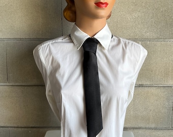 May include: A mannequin head with a vintage hairstyle, wearing a white button-down shirt and a black tie. The mannequin has a painted face with red lipstick and blue eye shadow. The background is a gray brick wall.