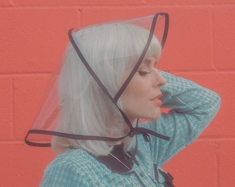 May include: A woman with white hair is wearing a clear plastic rain hat with a black trim. She is wearing a blue and white patterned sweater and has her hair pulled back.