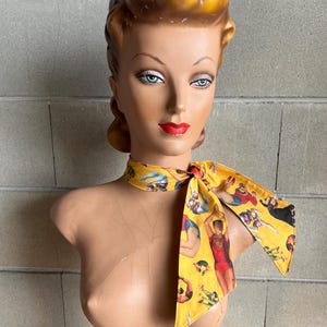 May include: A yellow scarf with a print of people in various circus poses. The scarf is tied around the neck of a mannequin with a blonde updo.