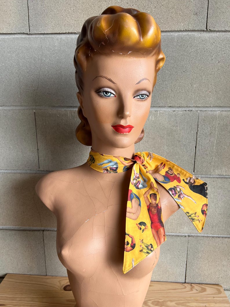 May include: A yellow scarf with a print of people in various circus poses. The scarf is tied around the neck of a mannequin with a blonde updo.