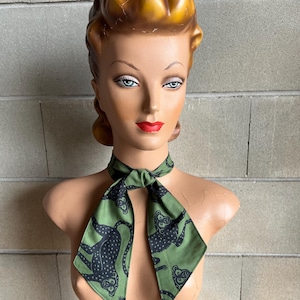 May include: A green scarf with a black monkey print. The scarf is tied around the neck of a mannequin.