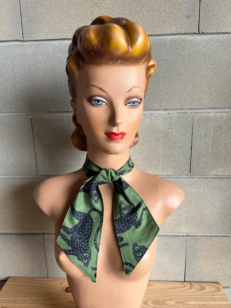 May include: A green scarf with a black monkey print. The scarf is tied around the neck of a mannequin.