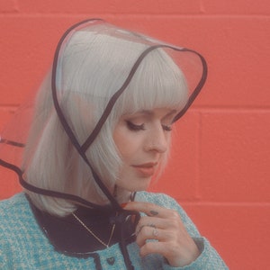 Op de afbeelding: Een vrouw met platinablond haar draagt een doorzichtige regenkap van plastic met een zwarte rand. Ze draagt een blauw en wit tweed jasje en heeft een zilveren ketting om.