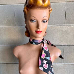 May include: A black scarf with a pattern of pink, blue, and gold hearts and wings. The scarf is tied around the neck of a mannequin with a blonde updo.