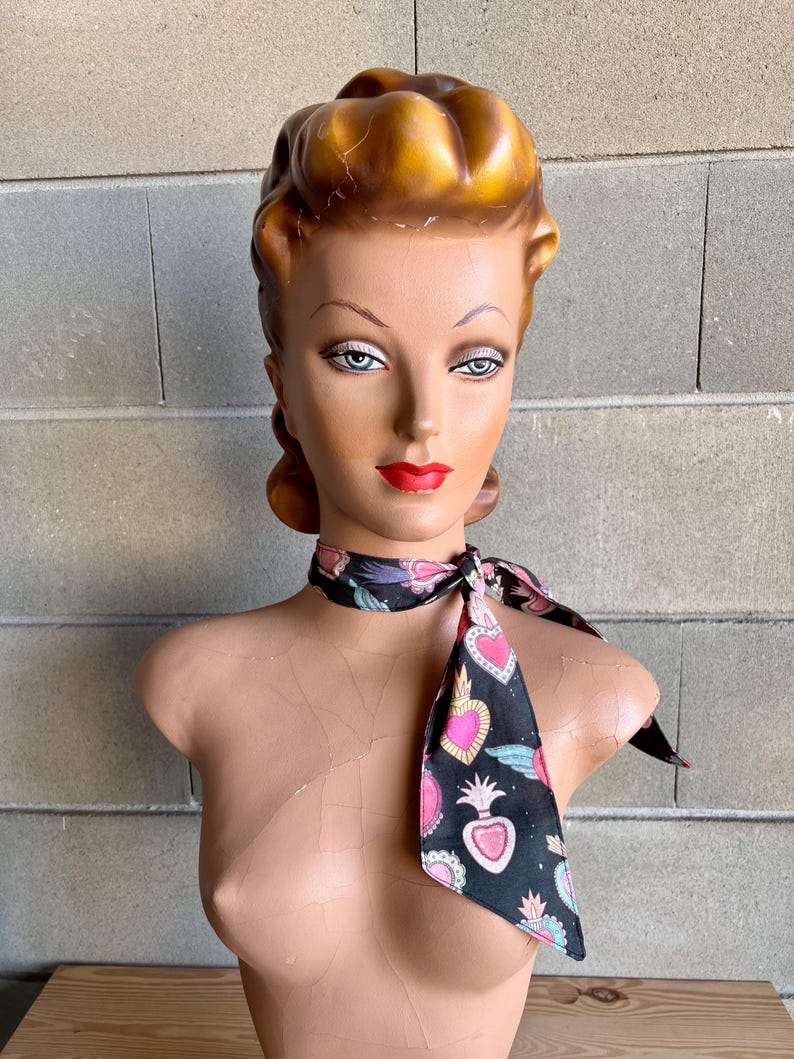 May include: A black scarf with a pattern of pink, blue, and gold hearts and wings. The scarf is tied around the neck of a mannequin with a blonde updo.