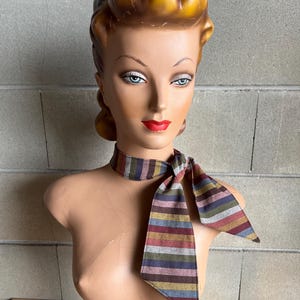 May include: A striped fabric neck scarf in shades of brown, gray, pink, and blue. The scarf is tied in a bow around the neck of a mannequin.