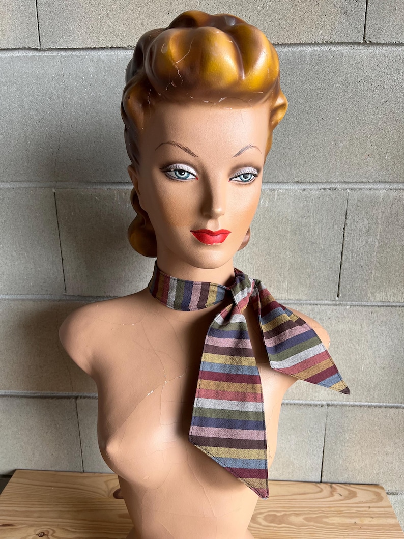 May include: A striped fabric neck scarf in shades of brown, gray, pink, and blue. The scarf is tied in a bow around the neck of a mannequin.