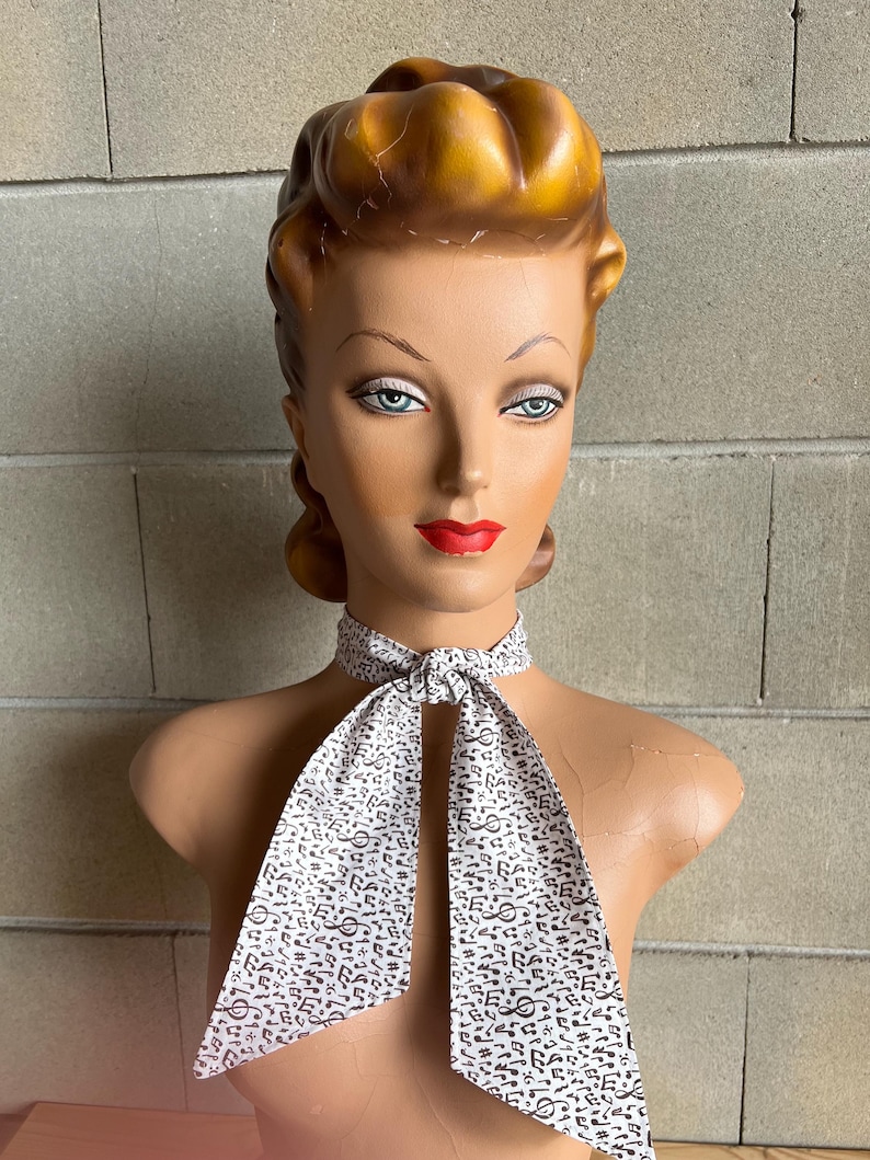 May include: A white necktie with a black musical note pattern. The necktie is tied around a mannequin's neck.
