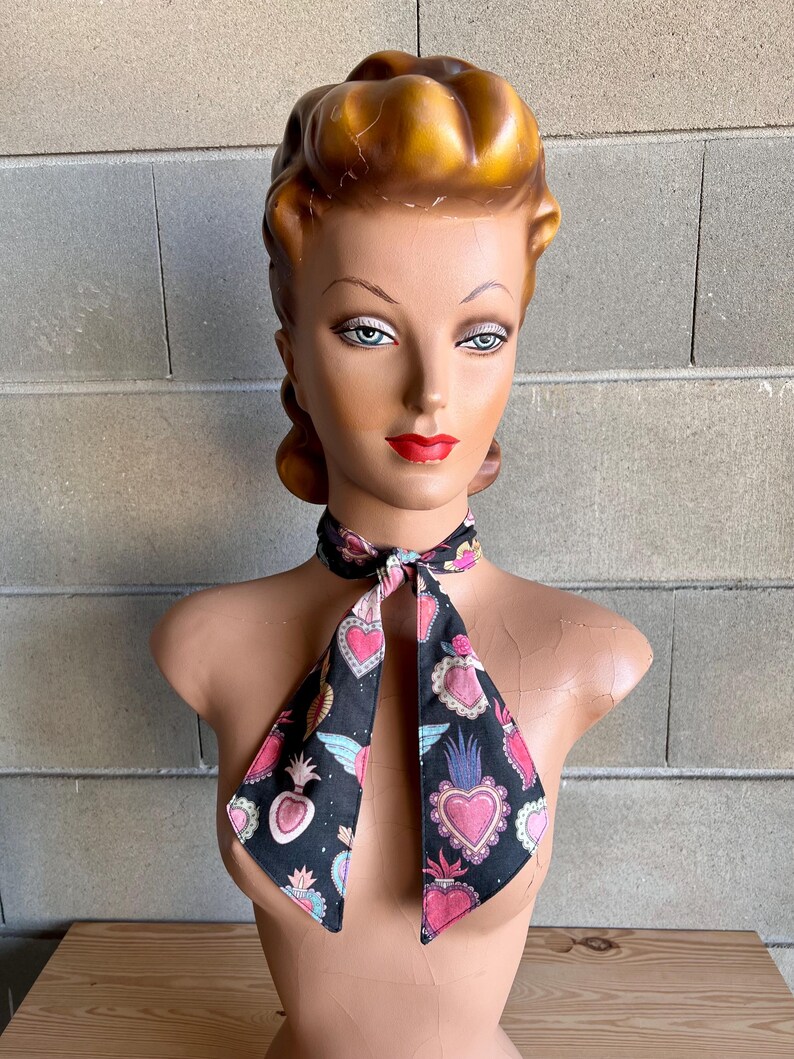 May include: A black necktie with a pattern of pink, blue, and white sacred hearts on a black background. The tie is tied in a bow around a mannequin's neck.