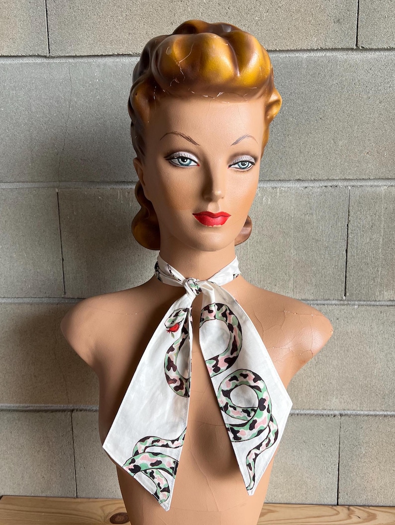 May include: A white silk scarf with a green and pink snake print. The scarf is tied around the neck of a mannequin.