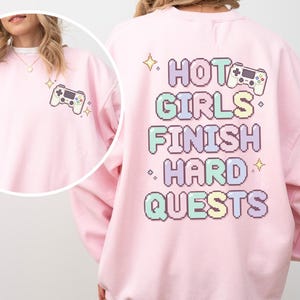 May include: A light pink crewneck sweatshirt featuring a small pixelated game controller graphic on the front left chest. The back displays the text HOT GIRLS FINISH HARD QUESTS in large, colorful pixelated letters with a game controller and stars.