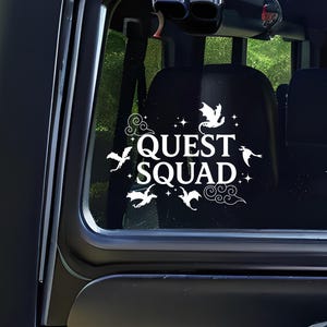 May include: White vinyl decal with the text "QUEST SQUAD" and a design of stylized dragons and clouds. The decal is on the back window of a black vehicle.