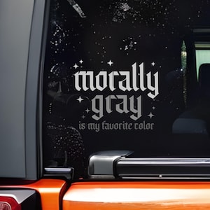 May include: A black car window decal with the words "morally gray is my favorite color" in a gothic font. The decal is white and has star accents. The decal is on the rear window of an orange vehicle.