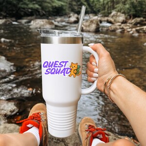 May include: White tumbler with a silver rim and handle, featuring the text "QUEST SQUAD" in purple with an orange cat illustration. The tumbler has a clear lid and a silver straw. The tumbler is held by a person.