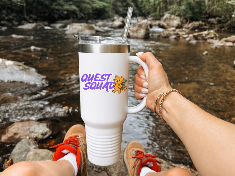 May include: White tumbler with a silver rim and handle, featuring the text "QUEST SQUAD" in purple with an orange cat illustration. The tumbler has a clear lid and a silver straw. The tumbler is held by a person.