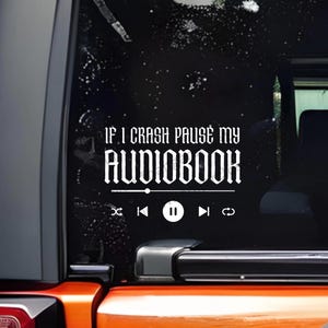 May include: White vinyl decal applied to a vehicle's rear window. The text reads "IF I CRASH PAUSE MY AUDIOBOOK" above a media player control bar with play, rewind, and fast forward icons. The background is black with white speckles.