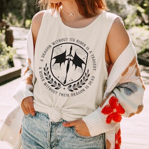 May include: Cream-colored tank top featuring a black and white dragon crest design with the text "A DRAGON WITHOUT ITS RIDER IS A TRAGEDY, A RIDER WITHOUT THEIR DRAGON IS DEAD." The top is knotted at the waist.