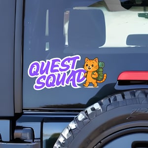 May include: A white and purple "Quest Squad" sticker featuring a cartoon orange cat with a green backpack. The sticker is affixed to the rear window of a vehicle, with the tire and taillight visible.