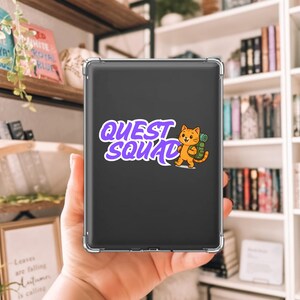 May include: A black tablet case with the words "QUEST SQUAD" in purple and white, accompanied by a cartoon orange cat with a backpack. The case is held in a person's hand, with a bookshelf in the background.