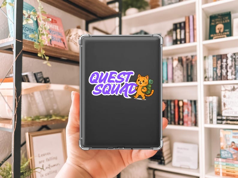 May include: A black tablet case with the words "QUEST SQUAD" in purple and white, accompanied by a cartoon orange cat with a backpack. The case is held in a person's hand, with a bookshelf in the background.