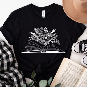 Puede incluir: Camiseta negra con un gr&aacute;fico blanco de un libro abierto con flores que salen de &eacute;l.