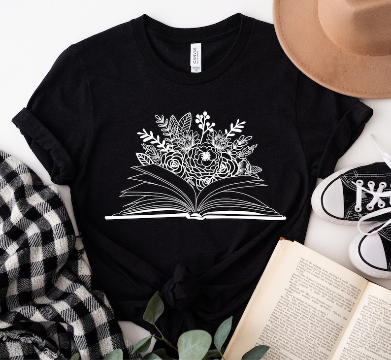 Puede incluir: Camiseta negra con un gr&aacute;fico blanco de un libro abierto con flores que salen de &eacute;l.