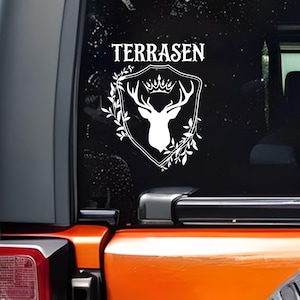 May include: White decal featuring a stag head with a crown inside a shield, surrounded by leaves. The word "TERRASEN" is above the shield. The decal is applied to the rear window of an orange vehicle.