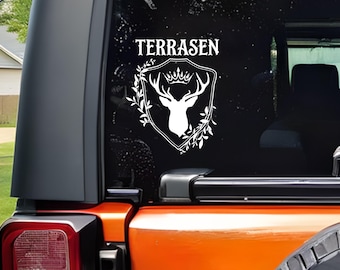 Puede incluir: Calcomanía blanca con la palabra "TERRASEN" sobre un escudo con un diseño de cabeza de ciervo, corona y corona de laurel. La calcomanía está en la ventana trasera de un vehículo naranja.