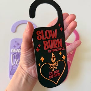 May include: Black door hanger with the text "SLOW BURN IN PROGRESS... DO NOT DISTURB" in red and orange. The design includes a candle inside a heart shape and starbursts. A purple and pink door hanger are also visible.
