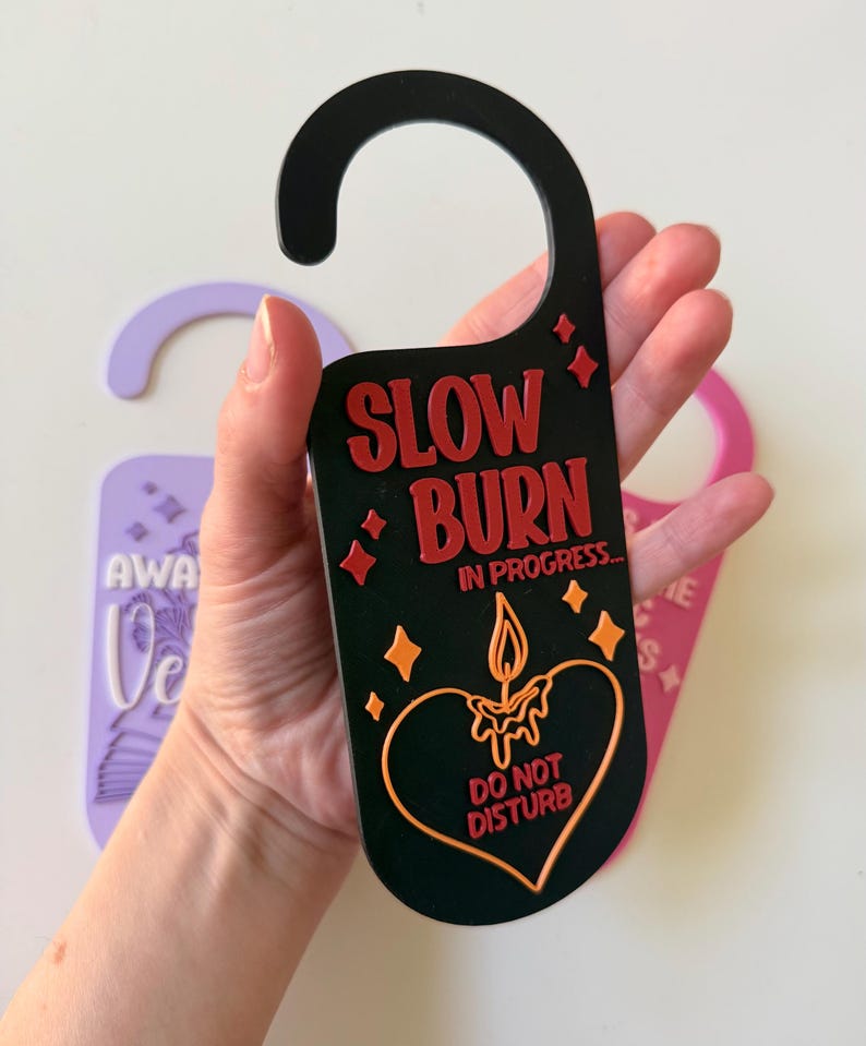 May include: Black door hanger with the text "SLOW BURN IN PROGRESS... DO NOT DISTURB" in red and orange. The design includes a candle inside a heart shape and starbursts. A purple and pink door hanger are also visible.