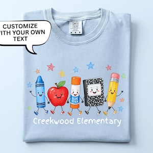 May include: A light blue t-shirt featuring a graphic of a blue crayon, a red apple, an orange glue stick, a black and white composition notebook, and a yellow pencil holding hands. The text reads "Creekwood Elementary" and "CUSTOMIZE WITH YOUR OWN TEXT".