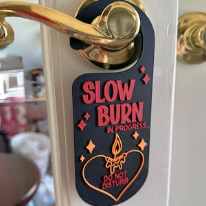 May include: A black door hanger with the words "SLOW BURN IN PROGRESS... DO NOT DISTURB" in red and orange. The design includes a candle inside a heart shape and star accents. The hanger is hanging on a door handle.