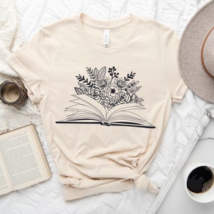 Puede incluir: Una camiseta beige con un gr&aacute;fico en blanco y negro de un libro abierto con flores que salen de &eacute;l.