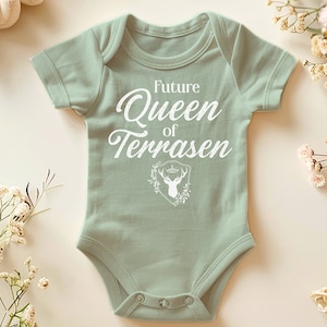 May include: A light green baby onesie with the text "Future Queen of Terrasen" in white. A white deer head with a crown is inside a shield with floral accents.