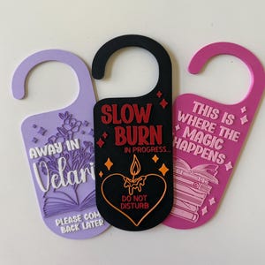 May include: Three decorative door hangers in purple, black, and pink. The purple hanger has floral and text details. The black hanger says "Slow Burn" and "Do Not Disturb". The pink hanger says "This is Where the Magic Happens".