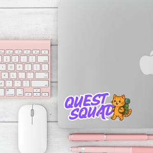 May include: A sticker with the text "QUEST SQUAD" in purple, featuring a cartoon orange cat with a backpack. The sticker is on a silver laptop, next to a pink keyboard, mouse, and pens.