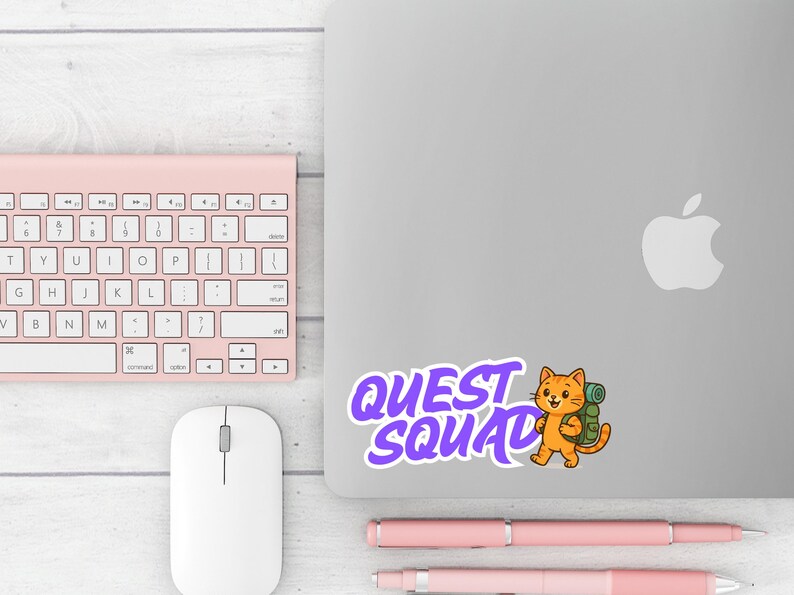 May include: A sticker with the text "QUEST SQUAD" in purple, featuring a cartoon orange cat with a backpack. The sticker is on a silver laptop, next to a pink keyboard, mouse, and pens.