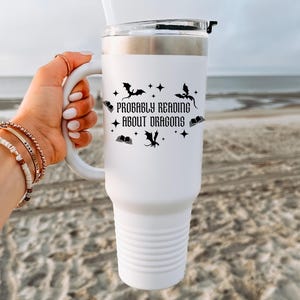 May include: White insulated travel mug with a handle, decorated with the text "Probably Reading About Dragons" and dragon and star motifs. The mug has a silver rim and a clear lid. The background is a beach scene.