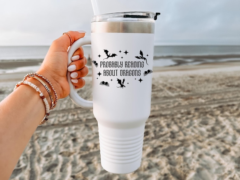 May include: White insulated travel mug with a handle, decorated with the text "Probably Reading About Dragons" and dragon and star motifs. The mug has a silver rim and a clear lid. The background is a beach scene.