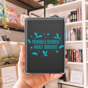 May include: A black tablet case featuring a teal dragon and star design, with the text "Probably Reading About Dragons". The case is held in front of a bookshelf filled with books.