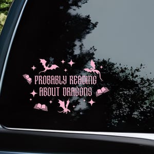 May include: A pink vinyl car sticker featuring the phrase "Probably Reading About Dragons". The design includes dragon silhouettes, open books, and stars. Suitable for application to a car window.