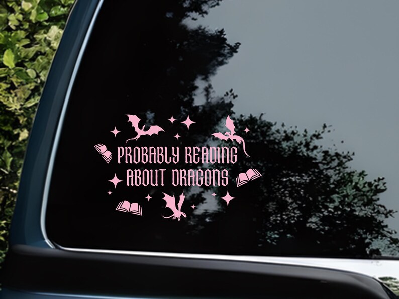 May include: A pink vinyl car sticker featuring the phrase "Probably Reading About Dragons". The design includes dragon silhouettes, open books, and stars. Suitable for application to a car window.