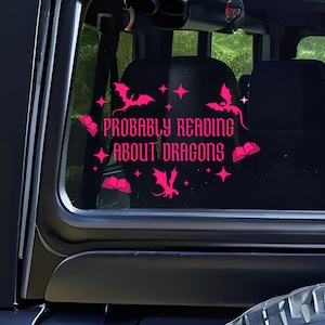 May include: A pink vinyl decal on a vehicle window. The decal reads "PROBABLY READING ABOUT DRAGONS" in a decorative font, with dragon and book illustrations. The interior of the vehicle is visible in the background.