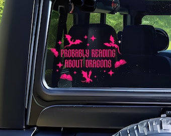 May include: A pink vinyl decal on a vehicle window. The decal features the phrase "PROBABLY READING ABOUT DRAGONS" in a stylized font, along with dragon and book illustrations. The background shows the interior of the vehicle.