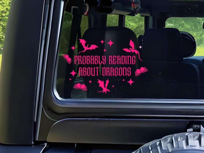 May include: A pink vinyl decal on a vehicle window. The decal reads "PROBABLY READING ABOUT DRAGONS" in a decorative font, with dragon and book illustrations. The interior of the vehicle is visible in the background.