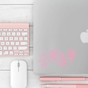 Op de afbeelding: Een werkpleksc&egrave;ne met een roze draadloos toetsenbord, muis en pennen. Een zilveren laptop toont roze driehoekige stickers met tekst. De items zijn gerangschikt op een licht houtnerfoppervlak.