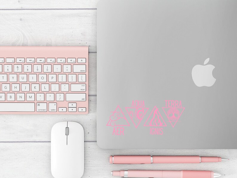 Op de afbeelding: Een werkpleksc&egrave;ne met een roze draadloos toetsenbord, muis en pennen. Een zilveren laptop toont roze driehoekige stickers met tekst. De items zijn gerangschikt op een licht houtnerfoppervlak.